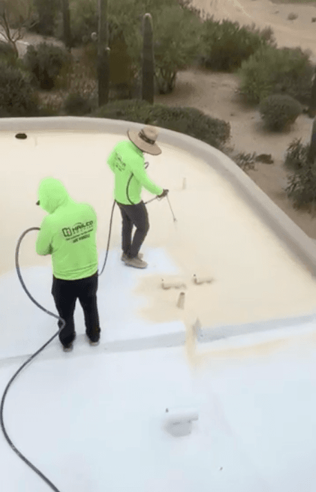 Two HailCo Roofing crew members in green hi-vis shirts rolling a bright white elastomeric coating over the new foam roof, saguaros and desert beyond