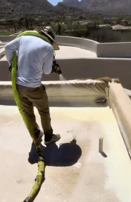 HailCo Roofing crew member pulling the spray-foam hose across a freshly coated parapet on the Desert Highlands estate, Pinnacle Peak desert beyond